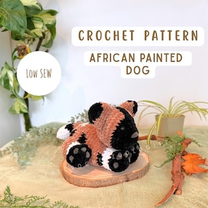 May include: A crocheted African painted dog plushie in brown, black, and white, resting on a wooden slice. The text "CROCHET PATTERN AFRICAN PAINTED DOG" is displayed above the plushie. The words "LOW SEW" are in a white circle.