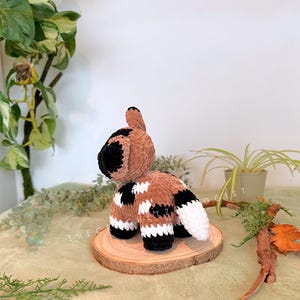 May include: A handmade crochet animal in brown, black, and white. The plush toy is positioned on a small wooden slice, with a white background and greenery. The toy has a tail and four legs.