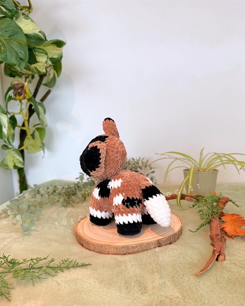 May include: A handmade crochet animal in brown, black, and white. The plush toy is positioned on a small wooden slice, with a white background and greenery. The toy has a tail and four legs.