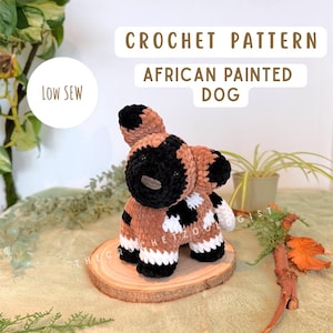 May include: A crocheted African painted dog in brown, black, and white. The soft toy is presented on a wooden slice. The image includes the text "CROCHET PATTERN AFRICAN PAINTED DOG" and "LOW SEW".