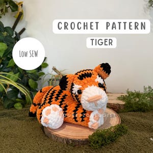 May include: A handmade crocheted tiger toy, featuring orange and black stripes, and white details on the face and paws. The text "CROCHET PATTERN TIGER" is displayed above the tiger. The toy is on a wooden slice, with the text "LOW SEW" in a white circle.