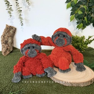 May include: Two handmade crochet orangutan plush toys. The plush toys are orange with gray faces, hands, and feet. One orangutan is sitting, and the other is reaching out. The toys are on a wooden slice on a green surface.