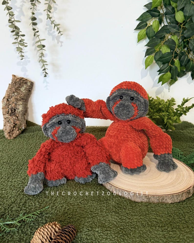 May include: Two handmade crochet orangutan plush toys. The plush toys are orange with gray faces, hands, and feet. One orangutan is sitting, and the other is reaching out. The toys are on a wooden slice on a green surface.