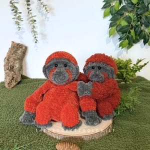 May include: Two handmade crochet monkey dolls. The monkeys are primarily red with gray faces, hands, and feet. They are sitting on a wooden slice, with the text "THE CROCHET ZOOOLOGIST" visible. The background is a green fabric.