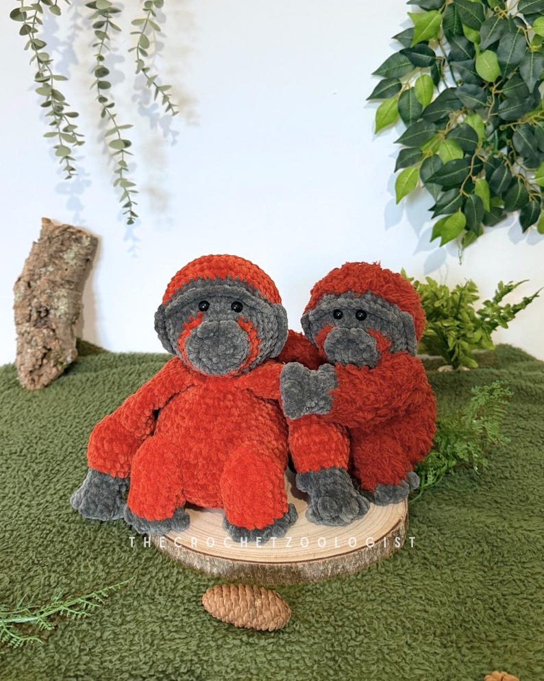 May include: Two handmade crochet monkey dolls. The monkeys are primarily red with gray faces, hands, and feet. They are sitting on a wooden slice, with the text "THE CROCHET ZOOOLOGIST" visible. The background is a green fabric.