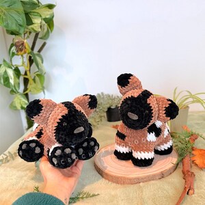 May include: Two handmade crochet African wild dog plushies. One is held in a hand, the other sits on a wooden slice. The plushies are brown, black, and white, with the word "CROCH" on the paw pads. The background includes greenery.