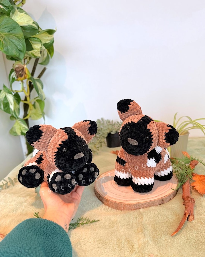 May include: Two handmade crochet African wild dog plushies. One is held in a hand, the other sits on a wooden slice. The plushies are brown, black, and white, with the word "CROCH" on the paw pads. The background includes greenery.