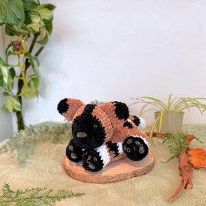 May include: A handmade crochet plush toy of a wild dog, featuring a black face and ears, brown and white body, and black paws. The plush is resting on a wooden slice. The text "THE CROCH ZOLO" is visible on the wooden slice.