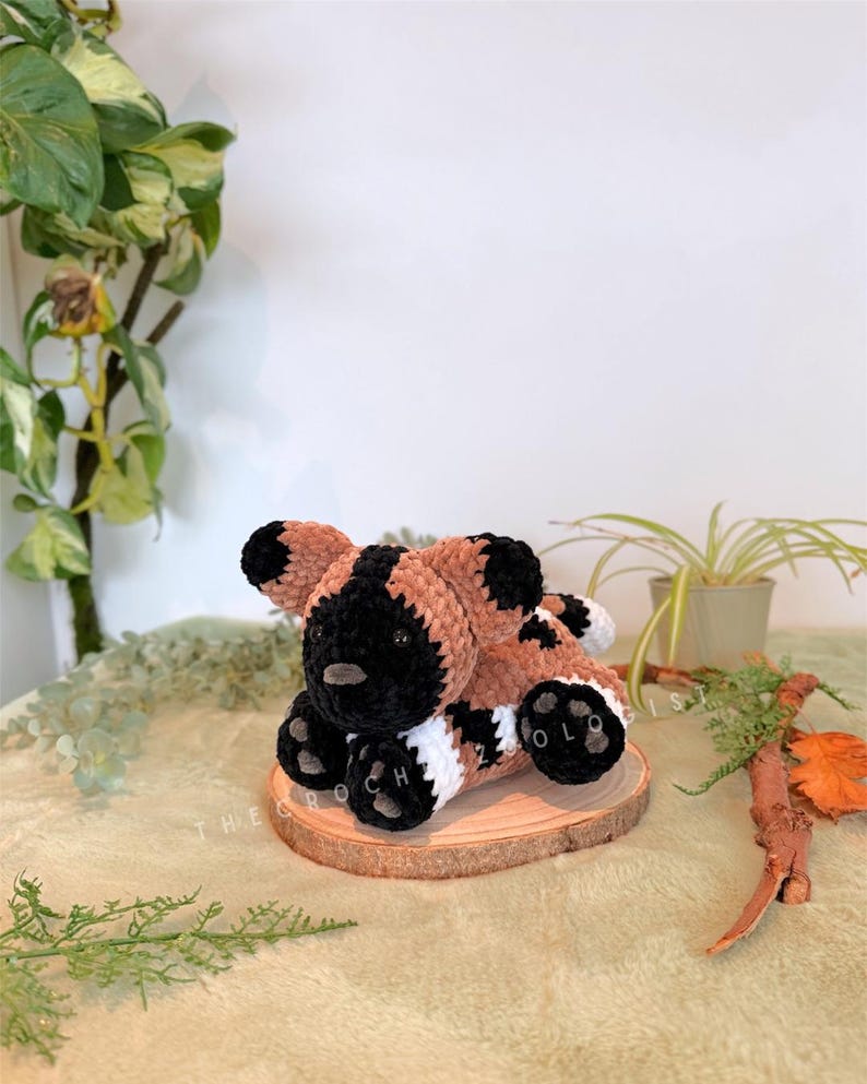 May include: A handmade crochet plush toy of a wild dog, featuring a black face and ears, brown and white body, and black paws. The plush is resting on a wooden slice. The text "THE CROCH ZOLO" is visible on the wooden slice.