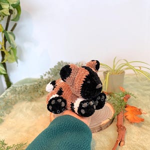 May include: A handmade crochet animal in brown, black, and white. The plush toy is sitting on a wooden slice. The text "THE CROC ZOO" is visible on the wooden slice.