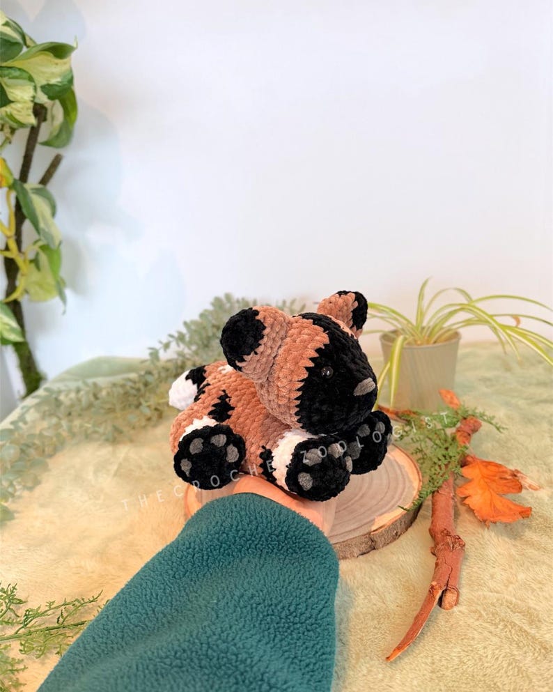 May include: A handmade crochet animal in brown, black, and white. The plush toy is sitting on a wooden slice. The text "THE CROC ZOO" is visible on the wooden slice.