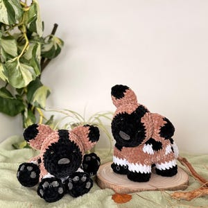 May include: Two handmade crochet African wild dog plushies. One is seated, the other standing on a wooden slice. The plushies feature black, tan, and white colors. The seated dog has black paws and a black nose. The standing dog has white and tan markings.