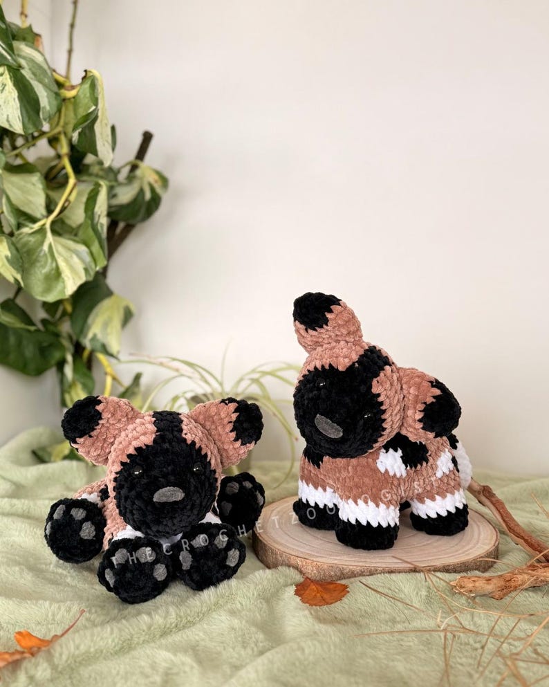 May include: Two handmade crochet African wild dog plushies. One is seated, the other standing on a wooden slice. The plushies feature black, tan, and white colors. The seated dog has black paws and a black nose. The standing dog has white and tan markings.