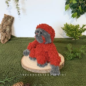 May include: A handmade, plush orangutan toy in shades of red and gray, sitting on a wooden slice. The crochet animal has a furry texture and is positioned against a green backdrop with foliage. The text "THECROCHETZOOLOGIST" is visible.