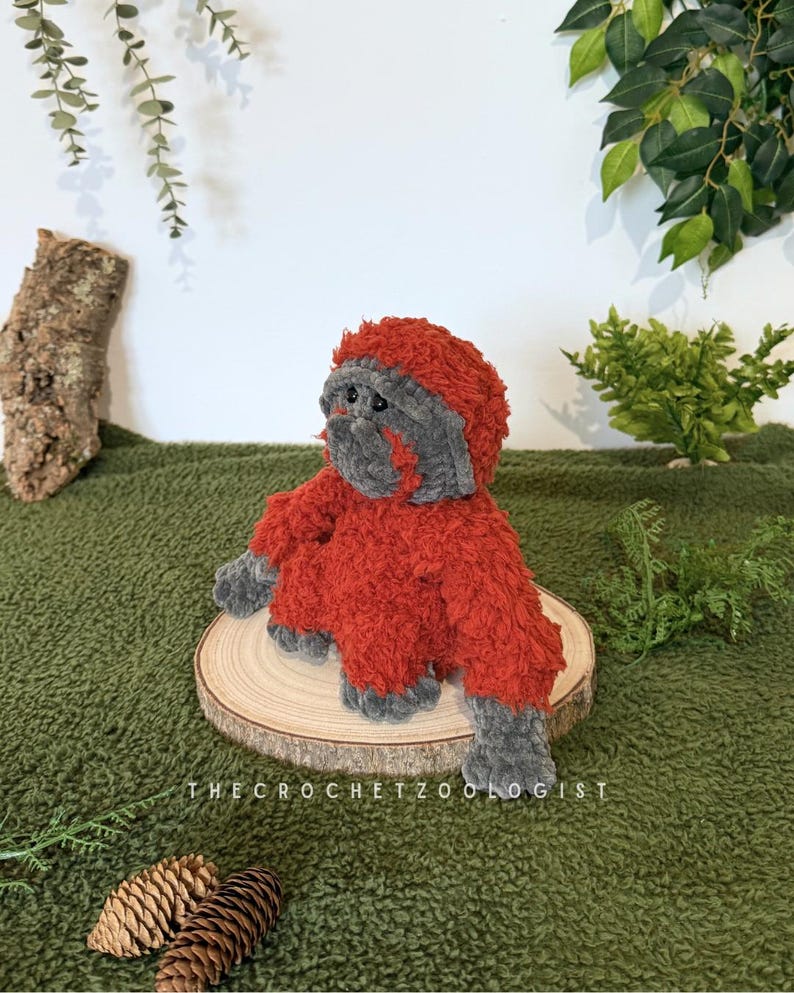 May include: A handmade, plush orangutan toy in shades of red and gray, sitting on a wooden slice. The crochet animal has a furry texture and is positioned against a green backdrop with foliage. The text "THECROCHETZOOLOGIST" is visible.