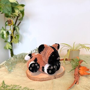 May include: A handmade plush toy African wild dog, in brown, black, and white, resting on a small wooden slice. The dog has black ears, a black muzzle, and black paws with white accents. The plush is soft and cuddly.
