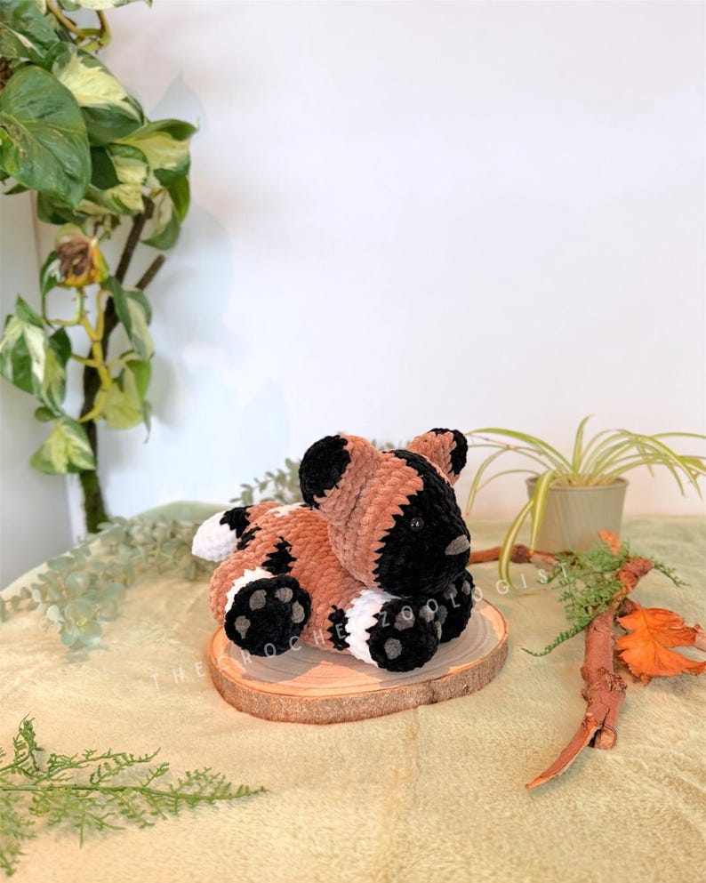 May include: A handmade plush toy African wild dog, in brown, black, and white, resting on a small wooden slice. The dog has black ears, a black muzzle, and black paws with white accents. The plush is soft and cuddly.