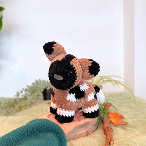 May include: A handmade crochet African wild dog plushie. The plushie features a black and tan color scheme with white accents. The dog is standing on a small wooden slice. The text "THE CROCHET GIST" is visible in the image.