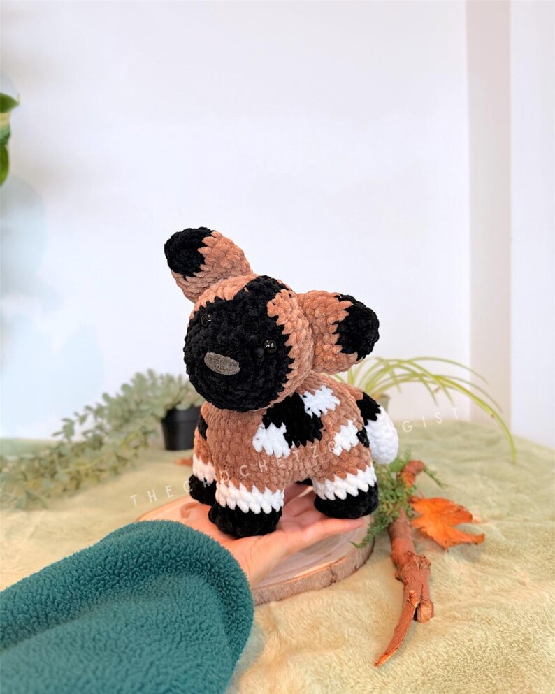 May include: A handmade crochet African wild dog plushie. The plushie features a black and tan color scheme with white accents. The dog is standing on a small wooden slice. The text "THE CROCHET GIST" is visible in the image.