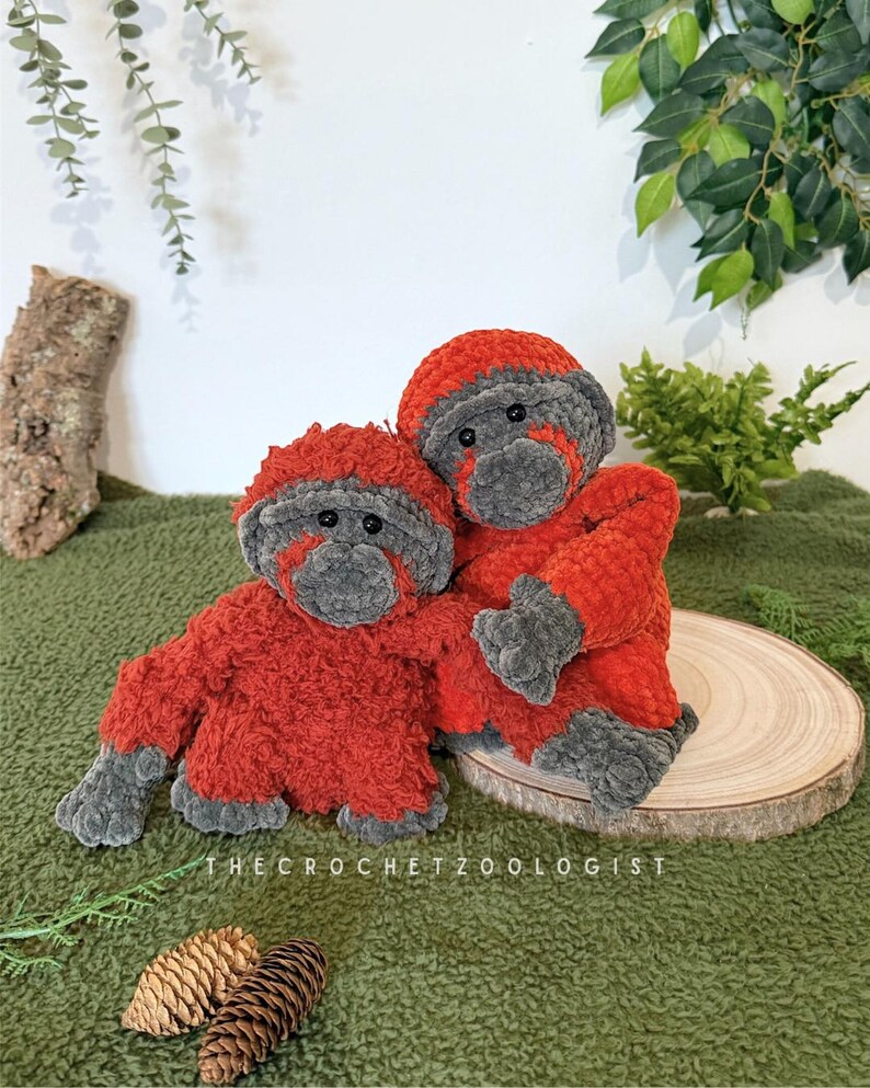 May include: Two handmade crochet orangutan plushies. The plushies are primarily red with gray accents on the face, hands, and feet. The text "THE CROCHET ZOOLOGIST" is visible at the bottom. The plushies are on a green surface.