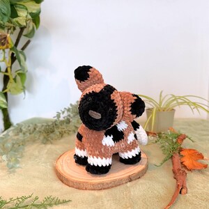 May include: A handmade crochet African wild dog plushie. The plushie features a brown body with black and white markings, black ears, and a black muzzle. It is sitting on a wooden slice, with greenery in the background.