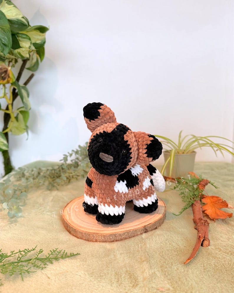 May include: A handmade crochet African wild dog plushie. The plushie features a brown body with black and white markings, black ears, and a black muzzle. It is sitting on a wooden slice, with greenery in the background.