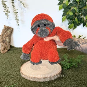 May include: A handmade crochet gorilla plush toy in shades of orange and gray. The gorilla is posed on a wooden slice, with the text "THE CROCHET ZOOLOGIST" visible. The plush has a soft, textured appearance.