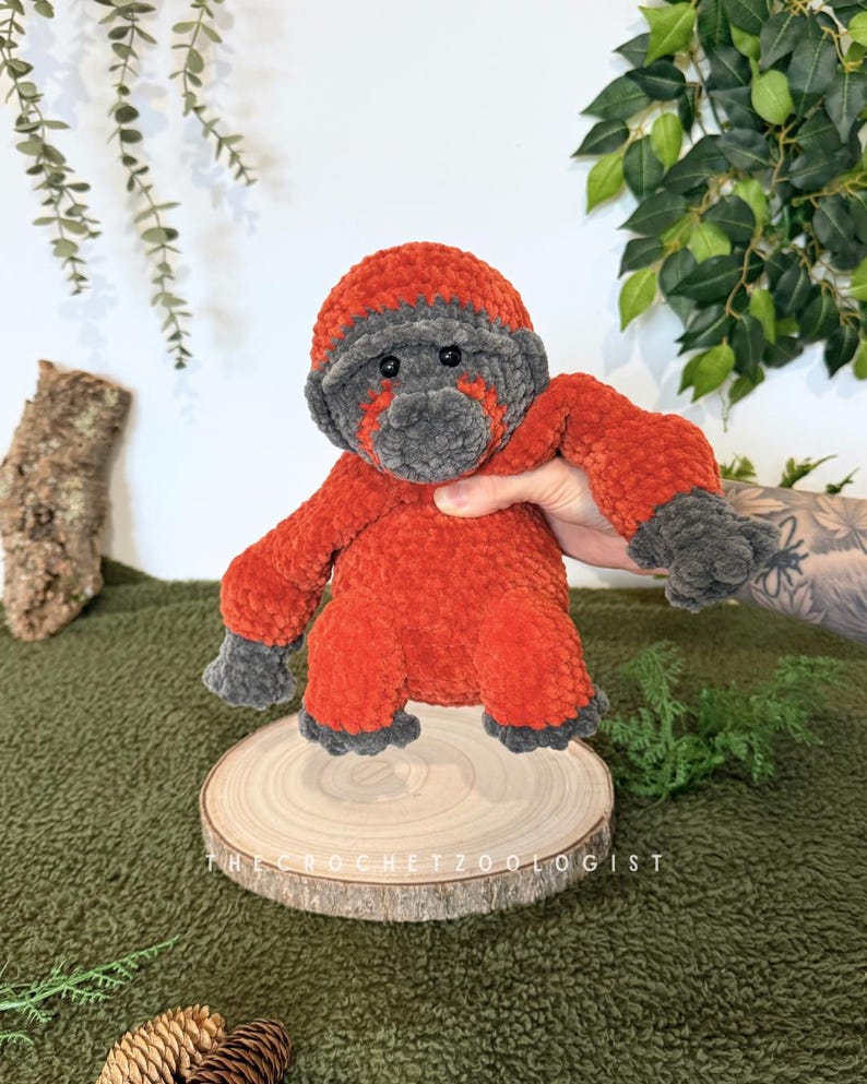 May include: A handmade crochet gorilla plush toy in shades of orange and gray. The gorilla is posed on a wooden slice, with the text "THE CROCHET ZOOLOGIST" visible. The plush has a soft, textured appearance.