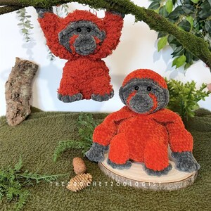 May include: Two plush orangutan toys. One hangs from a moss-covered branch, the other sits on a wooden slice. Both are orange with gray faces, hands, and feet. The text "THE CROCHET ZOOOLOGIST" is visible.