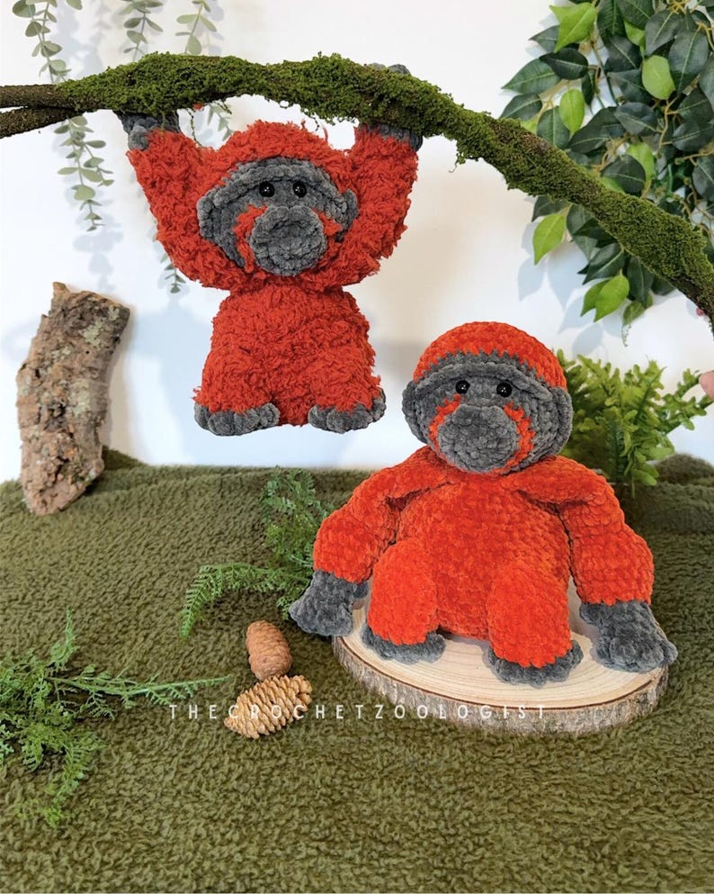 May include: Two plush orangutan toys. One hangs from a moss-covered branch, the other sits on a wooden slice. Both are orange with gray faces, hands, and feet. The text "THE CROCHET ZOOOLOGIST" is visible.