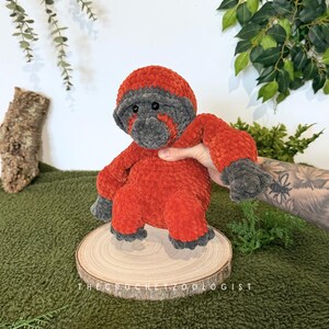 May include: A handmade, plush crochet gorilla in shades of orange and gray. The gorilla is seated on a wooden slice, with the text "THE CROCHET ZOOOLOGIST" visible. The background features greenery and a textured surface.