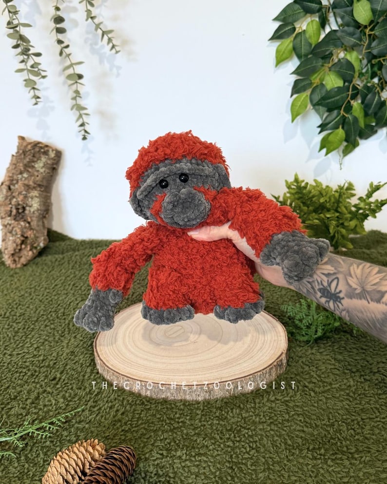 May include: A handmade crochet orangutan doll with red and gray fur. The plush toy is displayed on a wooden slice, with the text "THE CROCHETZOOLOGIST" visible. The background includes green foliage and a green textured surface.