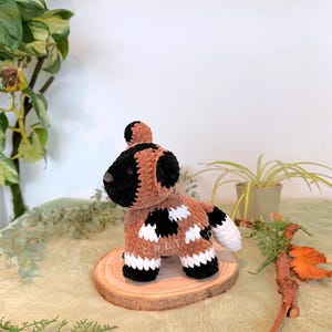 May include: A handmade crochet dog plushie in brown, black, and white, with a textured appearance. The dog is posed on a small wooden slice, showcasing its spotted pattern and fluffy tail. The plushie is a unique, handcrafted item.