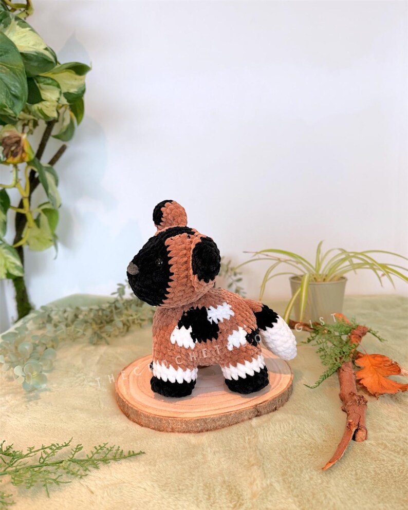 May include: A handmade crochet dog plushie in brown, black, and white, with a textured appearance. The dog is posed on a small wooden slice, showcasing its spotted pattern and fluffy tail. The plushie is a unique, handcrafted item.
