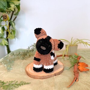 May include: A handmade crochet animal in brown, black, and white. The plush toy is a dog-like creature with a round head, black ears, and a black nose. It stands on a small wooden slice.