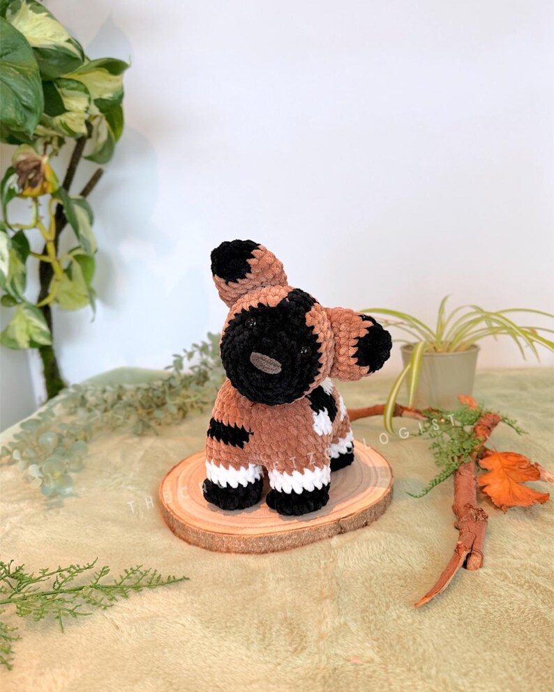 May include: A handmade crochet animal in brown, black, and white. The plush toy is a dog-like creature with a round head, black ears, and a black nose. It stands on a small wooden slice.