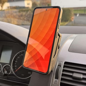 May include: A smartphone with an orange and red gradient screen is mounted in a car. The phone is in a black case and is held by a tan phone mount attached to the dashboard. The dashboard is gray.