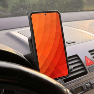 May include: A black smartphone in a car mount, displaying an orange and red gradient design. The phone is mounted on a car dashboard, near the steering wheel and air vents. The emergency flasher button is visible.