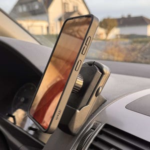 May include: A black smartphone in a protective case is mounted on a car dashboard using a phone holder. The phone's screen reflects the outside environment. The phone holder is black and silver.