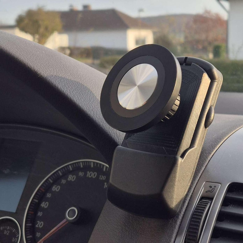 May include: A black and silver car phone mount attached to a car's dashboard. The mount features a circular magnetic head with a silver center and a black outer ring. The mount is designed to hold a phone securely while driving.