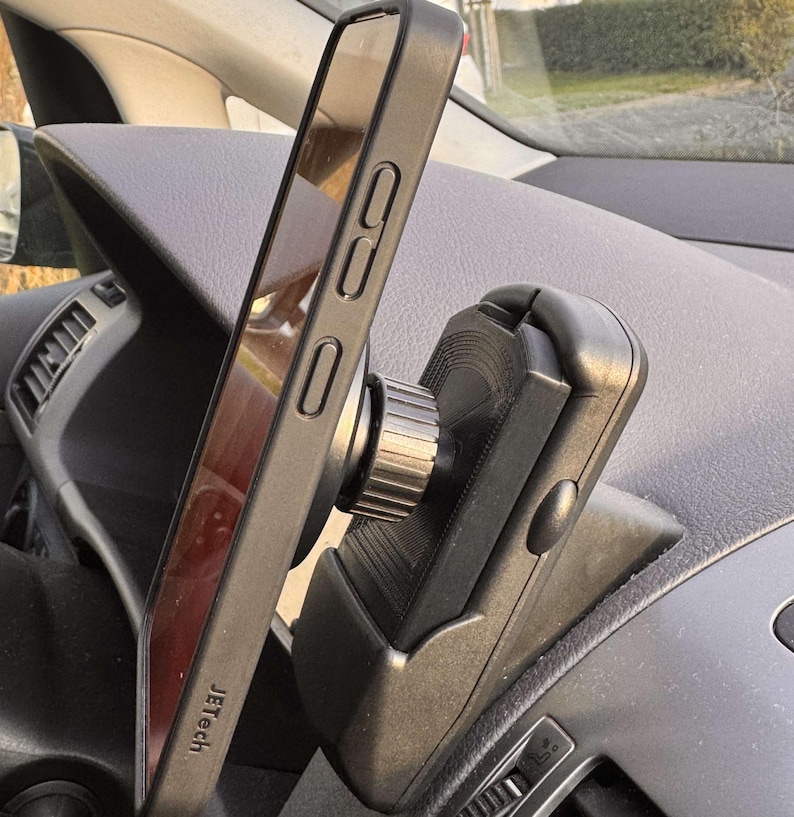 May include: Black smartphone in a protective case mounted on a car dashboard phone holder. The phone is angled, displaying the screen. The phone holder is black and has a rotating knob. The brand name "Jetech" is visible on the phone case.