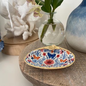 Puede incluir: Un plato decorativo en forma de concha de mar con un borde dorado y un patrón floral con mariposas, flores y hojas en tonos azules, rojos, naranjas y blancos. El plato está sobre un soporte de madera.