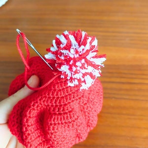May include: A red crocheted hat with a white and red pom-pom on top. A needle and thread are being used to attach the pom-pom to the hat.