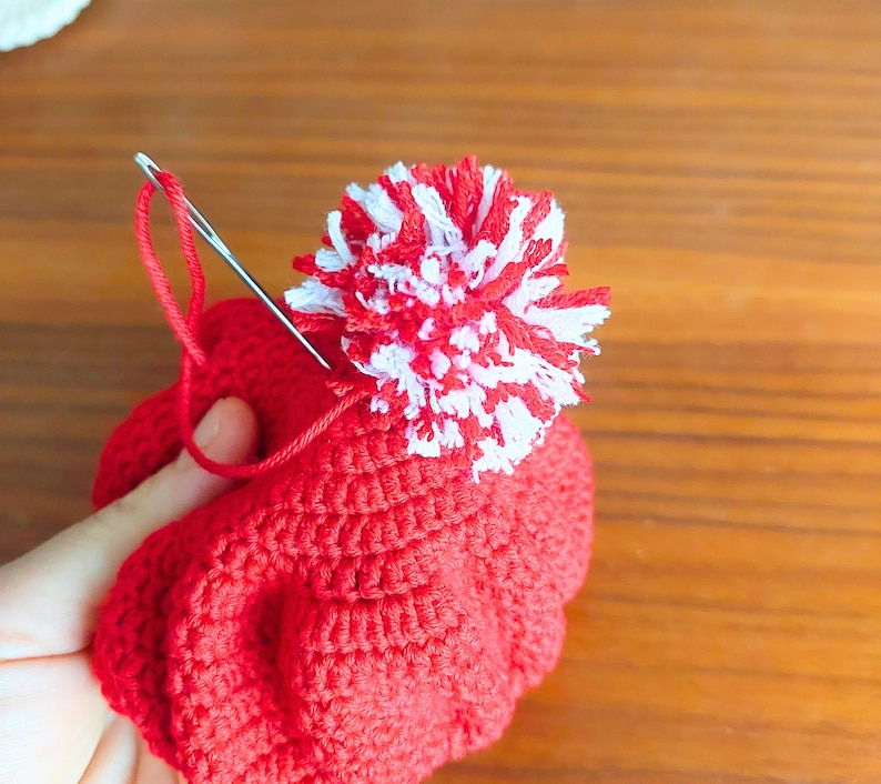 May include: A red crocheted hat with a white and red pom-pom on top. A needle and thread are being used to attach the pom-pom to the hat.
