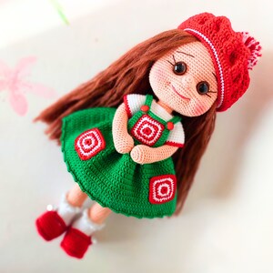 May include: A crocheted doll with long brown hair, wearing a green dress with red and white accents, and a red and white hat.