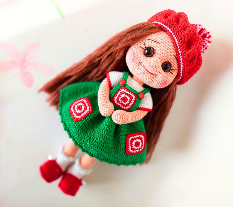 May include: A crocheted doll with long brown hair, wearing a green dress with red and white accents, and a red and white hat.