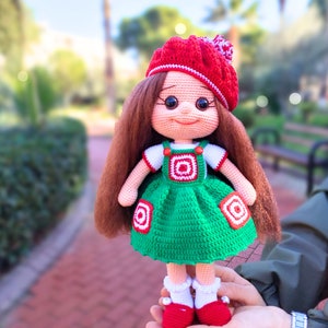 May include: A crocheted doll with long brown hair, wearing a red and white hat, a white shirt, and a green dress with red and white accents. The doll is holding a red and white ball.