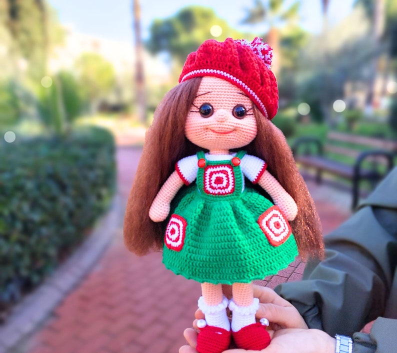 May include: A crocheted doll with long brown hair, wearing a red and white hat, a white shirt, and a green dress with red and white accents. The doll is holding a red and white ball.