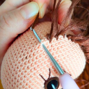 May include: A close-up of a crocheted doll head with brown yarn hair being sewn on. The doll has a pink crocheted face with a brown eye and a needle and thread are being used to attach the hair.