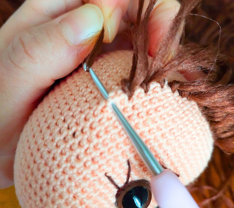 May include: A close-up of a crocheted doll head with brown yarn hair being sewn on. The doll has a pink crocheted face with a brown eye and a needle and thread are being used to attach the hair.